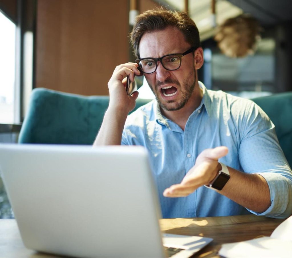 Stock photo of a man looking frustrated at a laptop computer and holding a smartphone to his ear