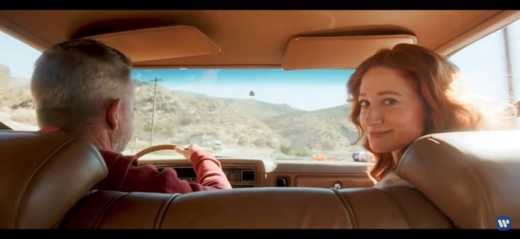 Two people in the front seat of a car as viewed from the back seat, and Bekka's character is glancing back toward the camera.