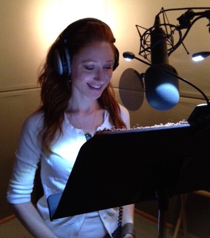 Bekka wearing headphones in a sound booth with a mic, reading from a music stand.