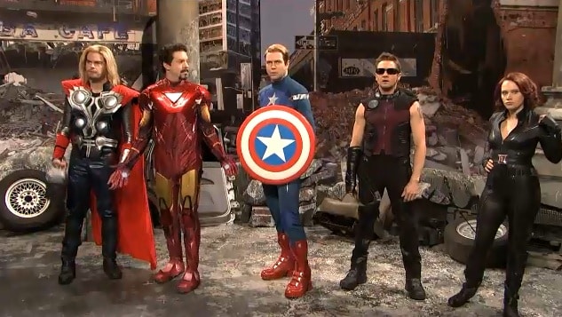 A wide shot showing Jeremy Renner as Hawkeye from Avengers, but in a Saturday Night Live comedy sketch with SNL cast members playing Thor, Iron Man, Captain America, and Black Widow, reproducing a battle scene from the first Avengers film.