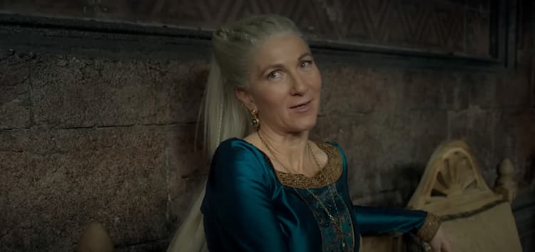 Close-up of Rhaenys Targaryen sitting against a stone wall and speaking to someone toward the camera.