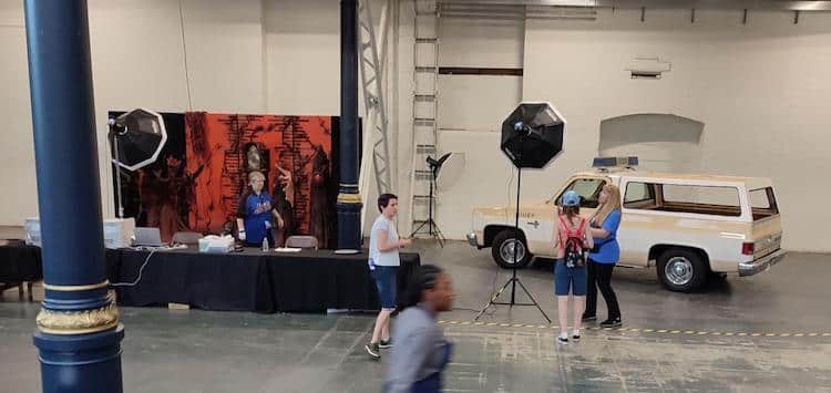 Wide image of a portion of a large convention space, the left side has a table on the left with a backdrop image from Stranger Things, and the right side as the sheriff's truck from Stranger Things. A few convention-goers are walking around the space.