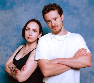 Lily K in a posed photo with Stranger Things actor Jospeh Quinn, partially back-to-back, both with arms crossed, with a blue backdrop.