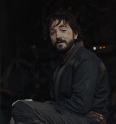 Cassian Andor crouches in the doorway of a spaceship, scavenging, but his head is raised with a slight smile as he's talking to someone out of frame.