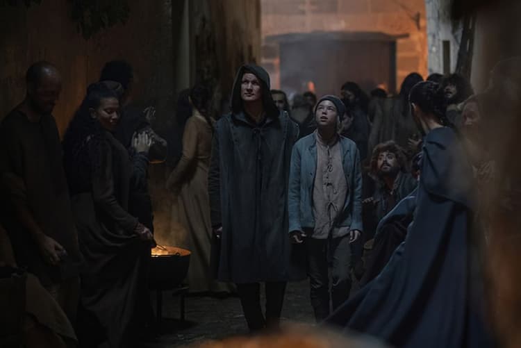 Daemon and Rhaenyra in the crowded streets of Flea Bottom. Daemon wears a hooded disguise and is walking with a focused look. Rhaenyra is dressed like a boy with a cap covering her hair, and she's looking up in fascination. 