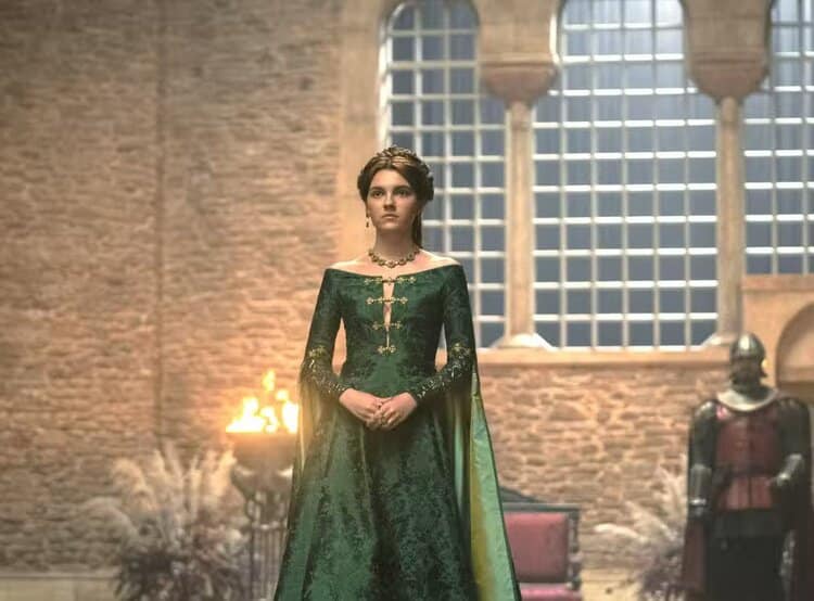Alicent stands alone in the brightly lit doorway to the throne room at the Red Keep, dressed in a gown of green brocade with gold trim, her hair in an elaborate up-do. Her expression appears confident, serious, and focused.