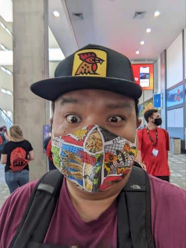 Selfie of Emmanuel Alejandro looking excited and wearing a comic strip print face mask while standing in the hallways of the Austin Convention Center while others are people walking around in the background. 