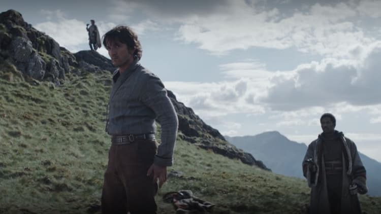 Cassian and other rebels stand on a grassy hillside during a cloudy day, one scouting with binoculars. Cassian is preparing to draw the blaster in his belt.