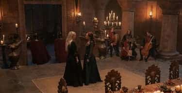 Alicent and Rhaenyra stand in an open area of a warmly-lit dining room, musicians playing their instruments in the background.