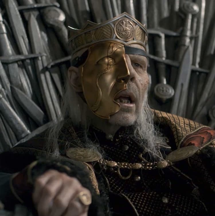 A closeup of King Viserys in his royal crown and robes sitting on the Iron Throne, his body resembling a gaunt corpse, and his face half-covered in a gold-colored half-mask.