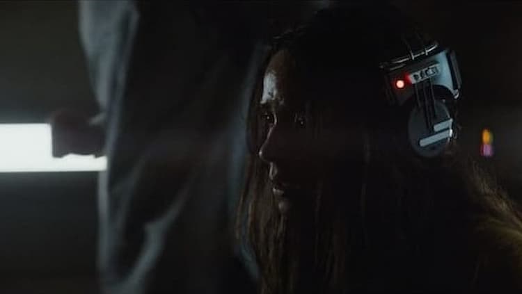Closeup of a distraught Bix in a dark room wearing a headphones-like torture device on her head.
