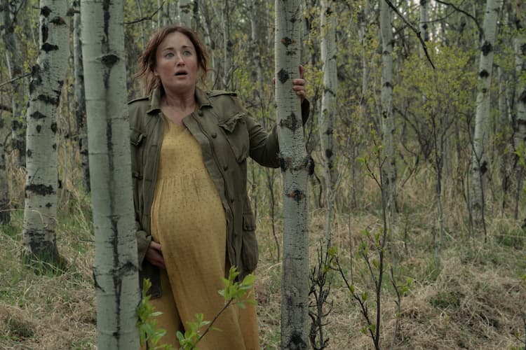 Anna, pregnant, looks worried standing in an outcrop of trees