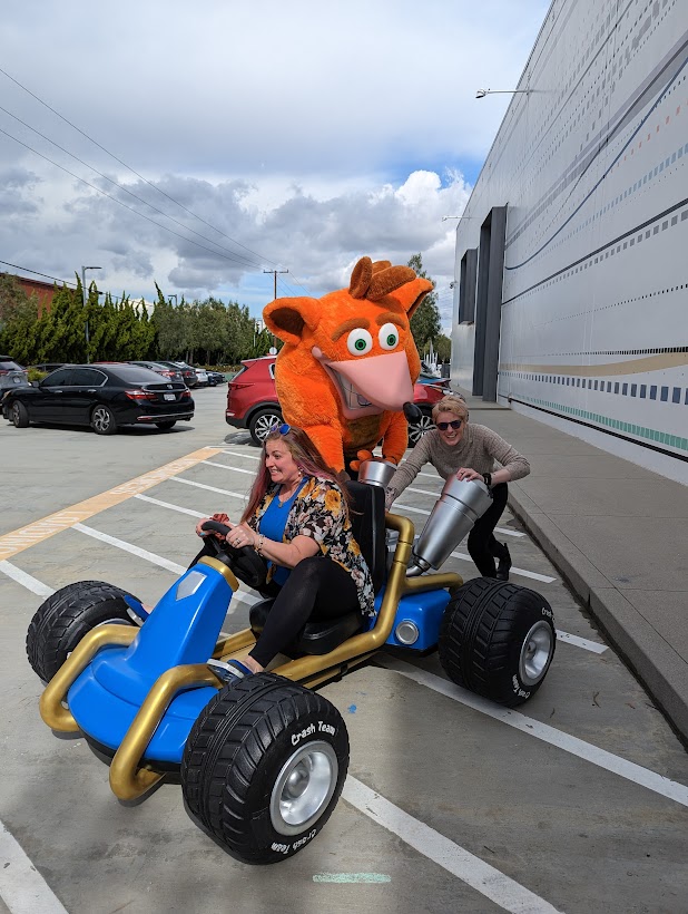 Michelle sits in a Crash Team Racing car as Erika and Crash Bandicoot push her along the parking lot.