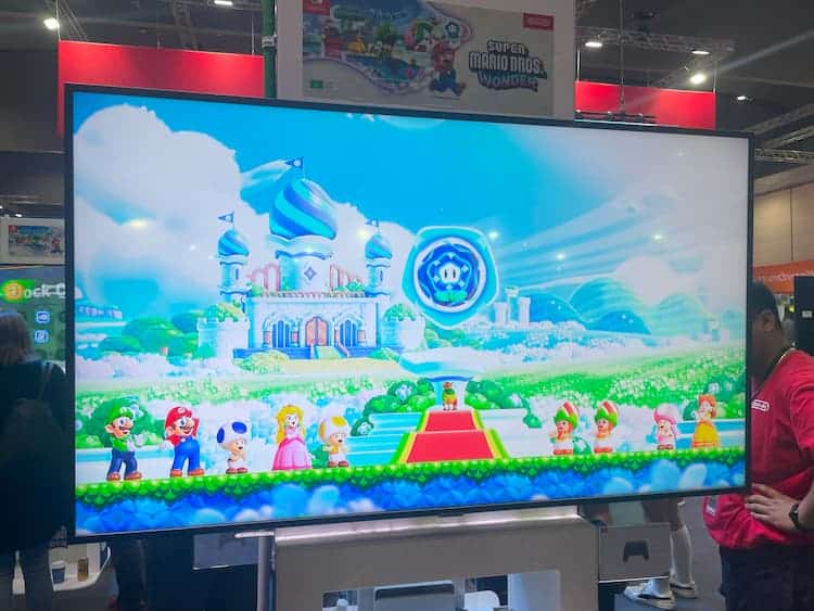 The intro cutscene of Super Mario Bros. Wonder is on the screen. Mario and his friends are standing with Prince Florian and some Poplins. A Wonder Flower is in the centre of the screen and a castle is in the background.