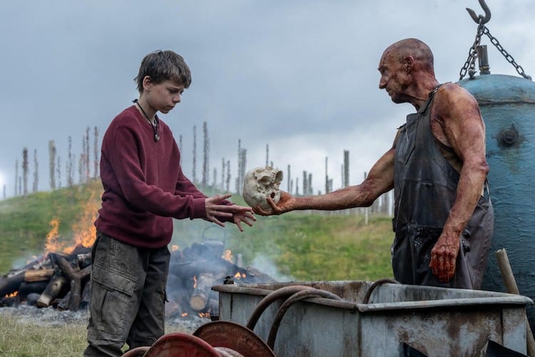 Kelson, wearing a leather apron, hands Spike a clean skull as a bonfire rages in the background.