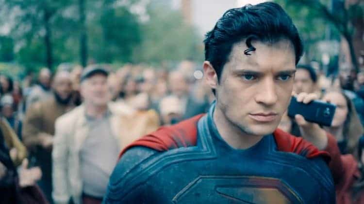 Superman is walking through a crowd. His suit is damaged. He has a depressed look on his face. The people around him are shouting and throwing stuff at him.